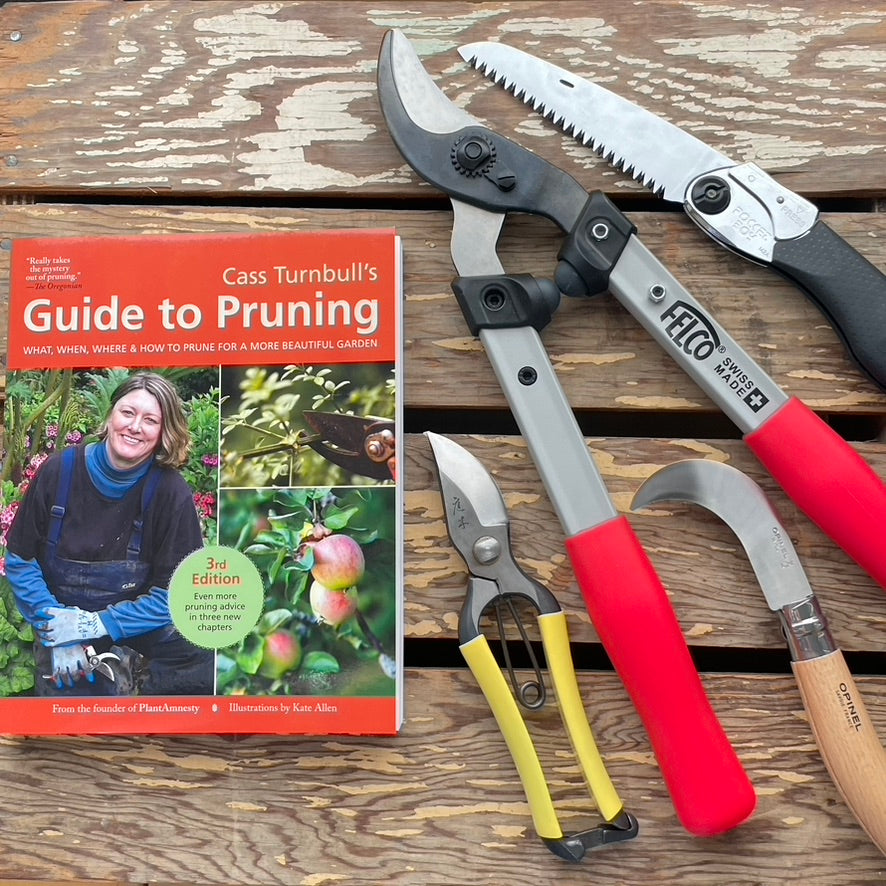 A gardening tool set arranged next to the book 'Cass Turnbull's Guide to Pruning, 3rd Edition.' The set includes pruning shears, a folding hand saw, hand pruner shears with yellow handles, a curved hand tool with a wooden handle, and a red-handled pruning tool.