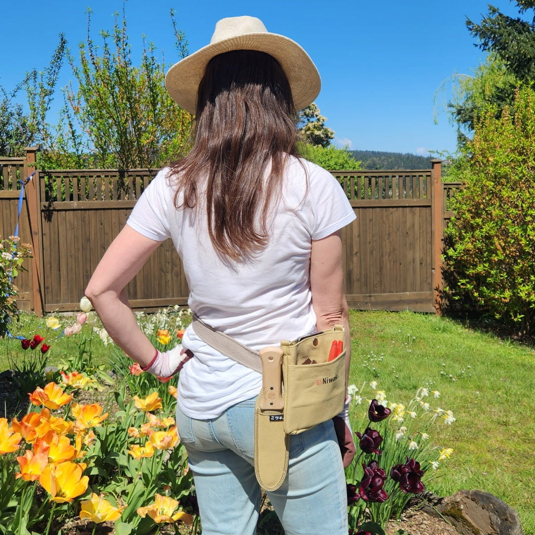 Person wearing a tool belt in a garden with flowers and a wooden fence in the background.