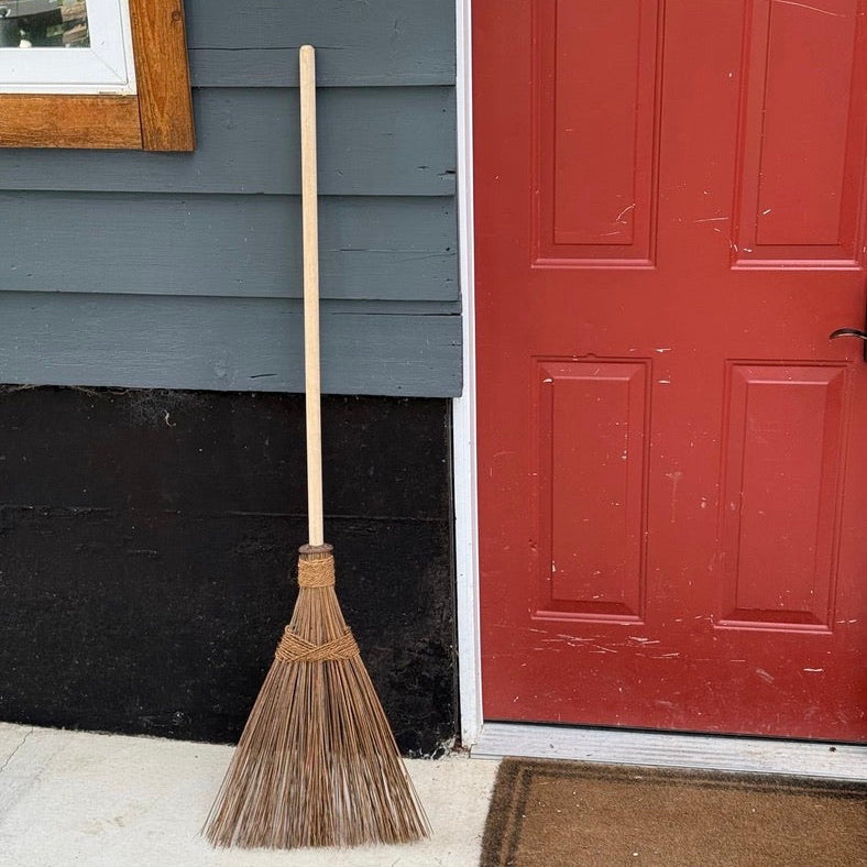A traditional straw broom leaning against a house exterior next to a red door and concrete porch.