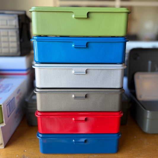 A stack of six plastic storage containers in assorted colors including green, blue, white, gray, red, and blue, displayed on a wooden surface.