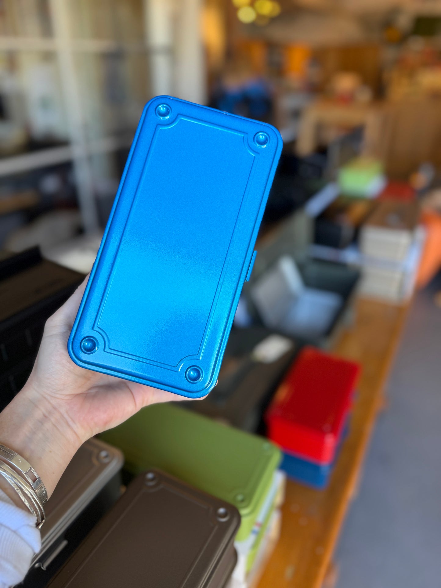 A hand holding a bright blue rectangular metal container with rounded corners in a blurred shop or storage room setting.