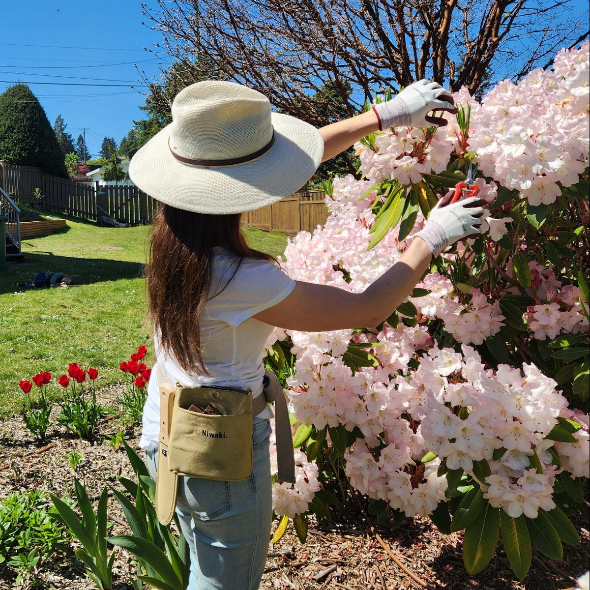 Person in a sunhat pruning pink flowers, with the tan Niwaki pouch at their hip.