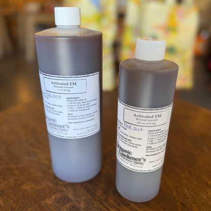 Two plastic bottles of Organic Gardener's Activated EM, one 1L and one 500 ml, standing on a wooden table with a blurred background.